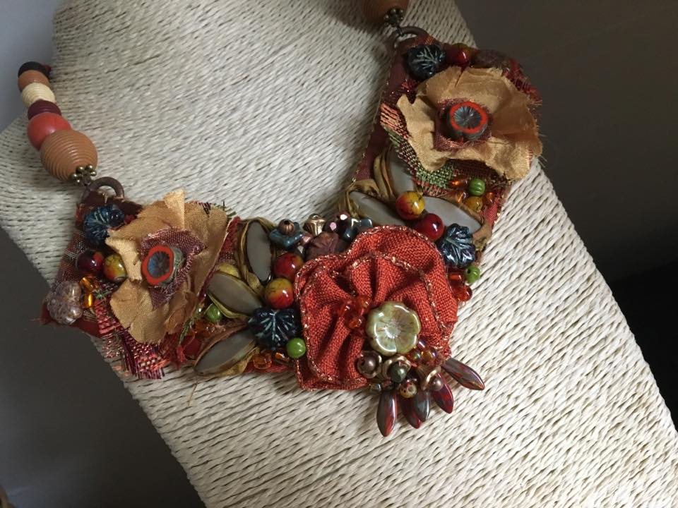 A statement collar necklace in rust and ochre fabric, set with ceramic flowers, glass beads and brass leaves.