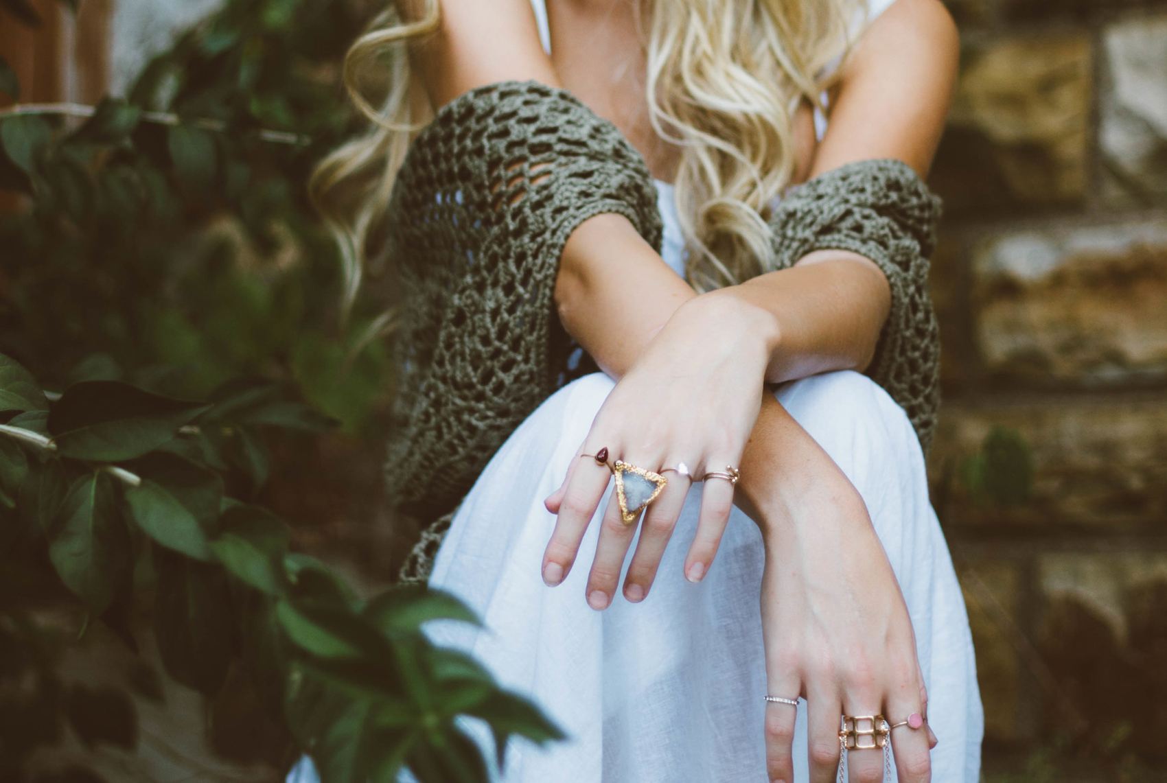 A woman sitting outdoors in a white dress and olive knit, wearing several handmade rings.